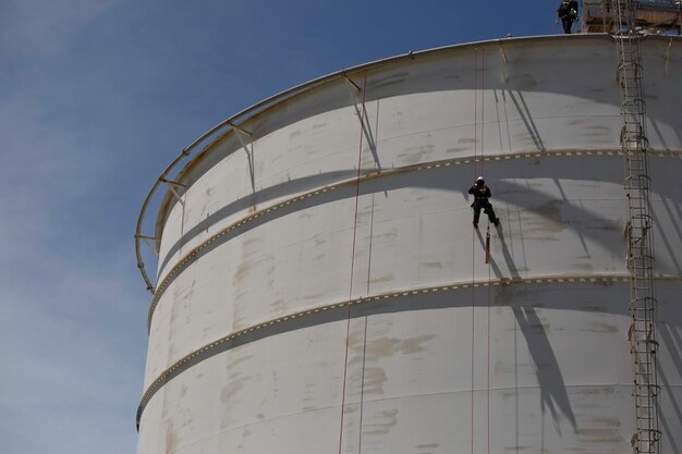 Работник-мужчина спускается по веревке на крыше резервуара, проверяет безопасность, толщина сварного шва резервуара для хранения газа
