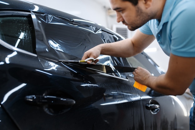 Il lavoratore maschio tiene il foglio di pellicola, l'installazione della colorazione dell'auto, il servizio di messa a punto meccanico che applica tinta vinilica sul finestrino del veicolo in garage, vetri oscurati per automobili