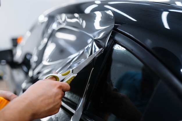 Male worker holds sheet of film, car tinting installation, tuning service. Mechanic applying vinyl tint on vehicle window in garage, tinted automobile glass