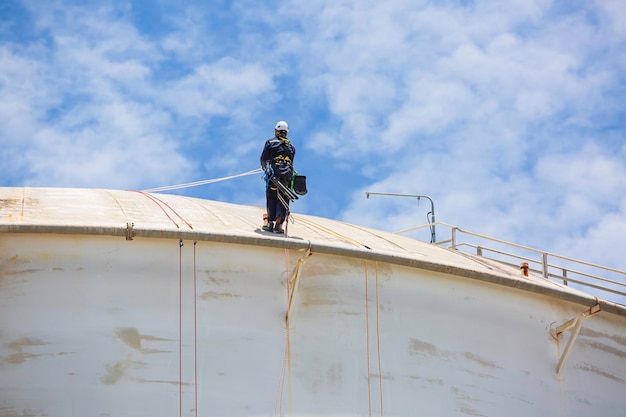 Ispezione del lavoratore di sesso maschile che indossa la prima linea di sicurezza della fune dell'imbracatura di sicurezza che lavora in un punto elevato sul cielo blu del gas sferico del tetto del serbatoio