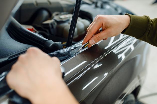 Male worker installs transparent protection film on car hood. Installation of coating that protects the paint of automobile from scratches. New vehicle in garage, tuning