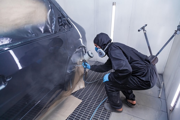 A male worker in jumpsuit and blue gloves paints with a spray gun a side part of the car body in black after being damaged at an accident Auto service industry professions