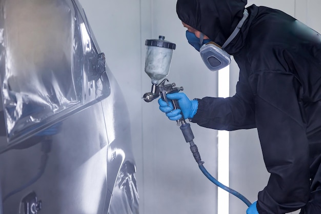 A male worker in jumpsuit and blue gloves paints with a spray gun a side part of the car body in black after being damaged at an accident Auto service industry professions