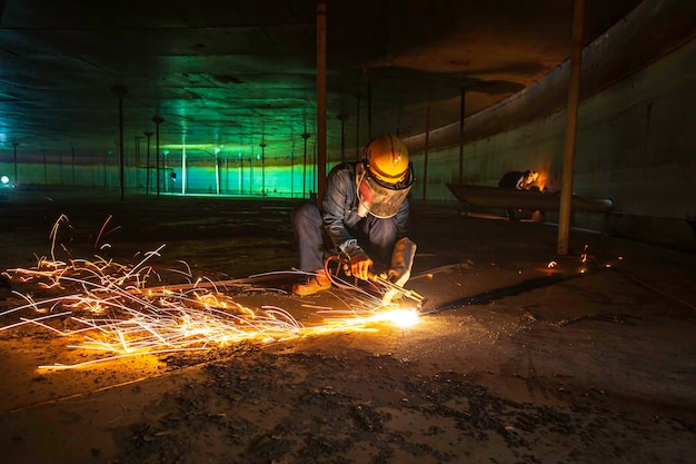 Рабочий-мужчина режет искру металла на стальной пластине дна резервуара со вспышкой режущего света крупным планом, носит защитные перчатки и маску в боковом замкнутом пространстве.