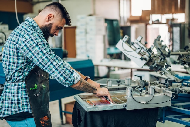 Male worker pressing ink on frame while using the printing\
machine in a workshop