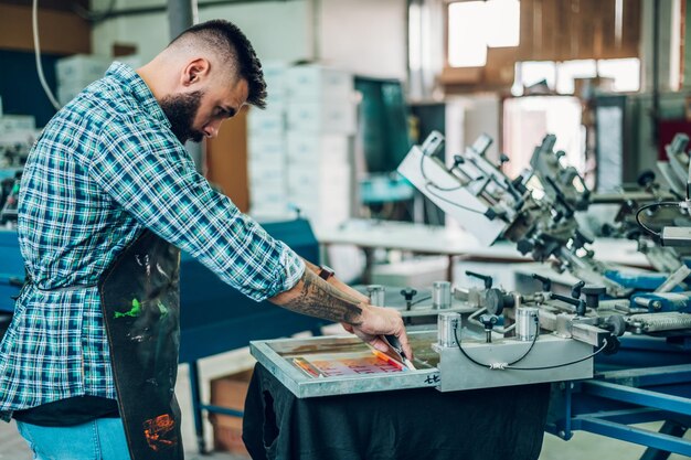 Работник-мужчина нажимает чернила на раму во время использования печатной машины в мастерской