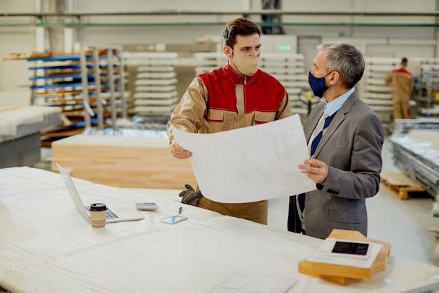 Male worker and project manager with face masks talking while examining plans at carpentry workshop