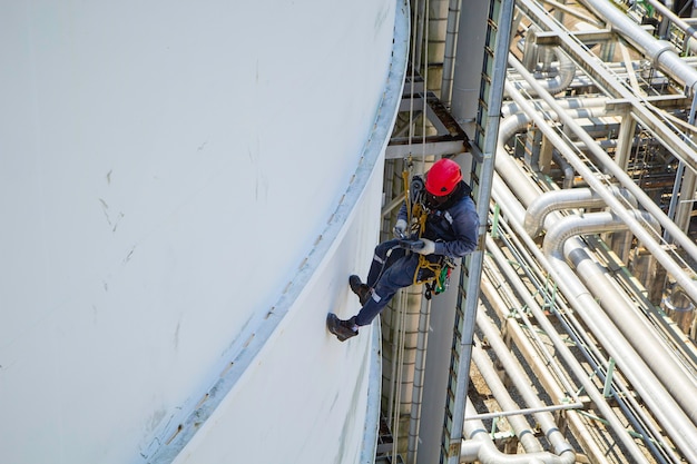 Male worker rope access height safety inspection of thickness\
storage oil and gas tank industry cloud blue sky.