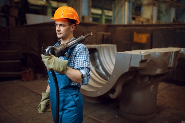 Il lavoratore maschio in uniforme e casco tiene il martello pneumatico pneumatico sulla fabbrica. industria metalmeccanica, produzione industriale di prodotti siderurgici