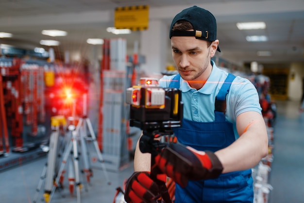 Male worker in uniform testing laser level on tripod in tool store