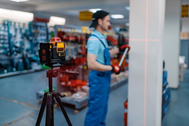 Male worker in uniform testing laser level on tripod in tool store