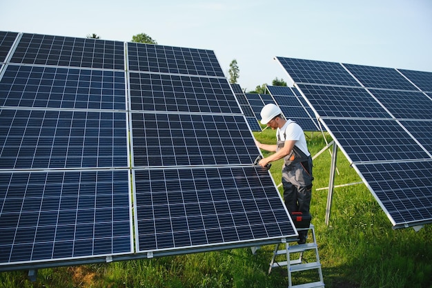 Male worker in uniform with a drill screwdriver in his hand on a stepladder near solar panels