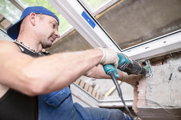 male worker uses electric perforator to drill hole in brick wall to install and fasten a PVC window