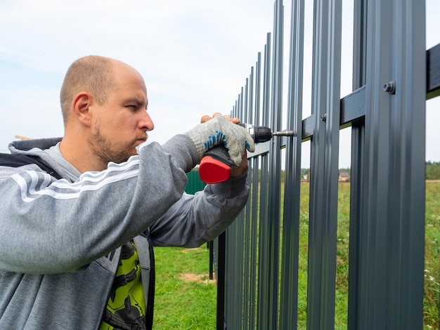 A male worker using an electric screwdriver twists a metal\
picket fence on the fence worker professional builder fencing\
security private territory