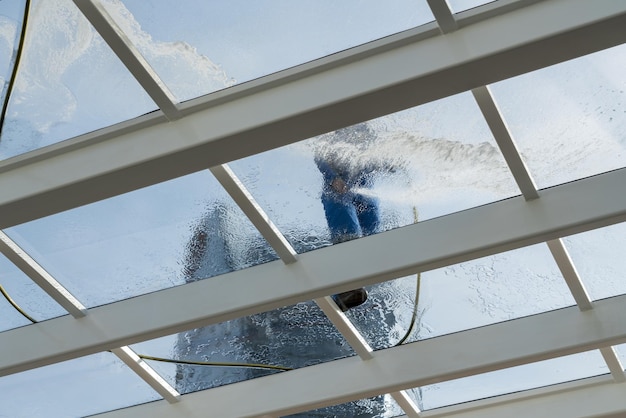 Male worker washing large expanse of glass roof over swimming pool with hose pipe