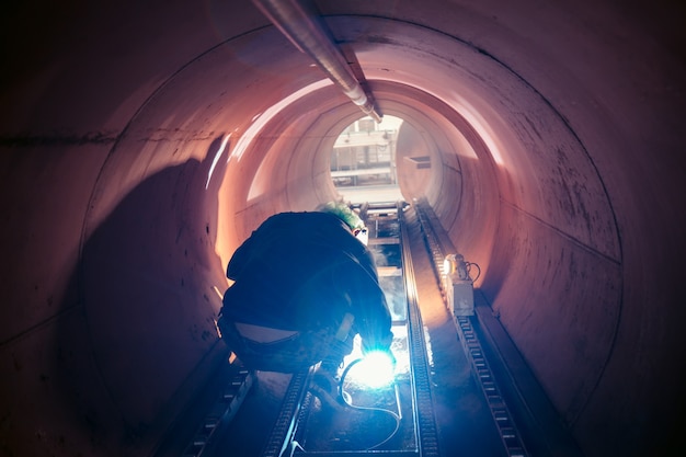 保護服を着用し、限られたスペース内で工業用建設の石油とガスまたは水平貯蔵タンクを修理溶接する男性労働者。