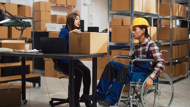 Male worker with impairment taking boxes off of shelves in storage room, organizing merchandise on racks. Young man in wheelchair talking to woman about business development and distribution.