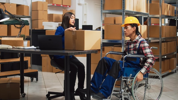 Foto lavoratore di sesso maschile con disabilità che preleva scatole dagli scaffali nel magazzino, organizzando la merce sugli scaffali. giovane in sedia a rotelle che parla con una donna dello sviluppo e della distribuzione del business.