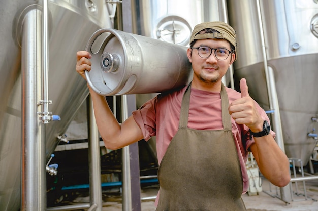 醸造所の中小企業とビール業界で金属製のビール樽を持つ男性労働者倉庫で彼の肩に鋼のバケツを持っているエプロンでハンサムなアジア人の笑顔
