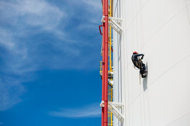 Male workers control swing rope down height tank rope access\
inspection of thickness pipeline and tank gas background blue\
sky.