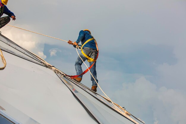 Рабочие-мужчины поднимаются по веревке на высоту безопасности, соединяясь с узлом ремней безопасности, врезающихся в купол масляного бака на строительной площадке крыши