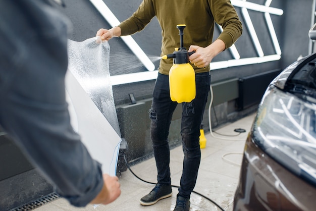 I lavoratori di sesso maschile bagnano il modello del parafango anteriore della pellicola di protezione dell'auto. installazione di rivestimento che protegge la vernice dell'automobile dai graffi. veicolo nuovo in garage, procedura di messa a punto