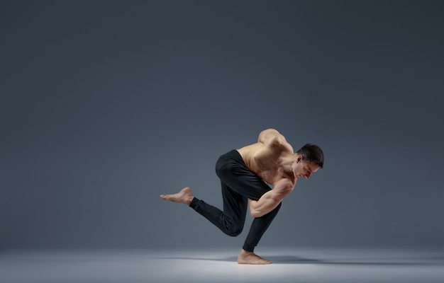 Male yoga keeps balanc in a difficult pose, grey background. Strong man doing yogi exercise, asana training, top concentration, healthy lifestyle