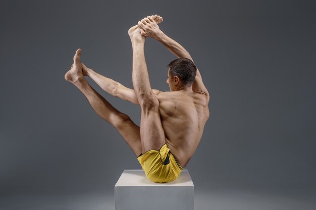 Male yoga sitting on the pedestal in studio, meditation position, grey wall Strong man doing yogi exercise, asana training, top concentration, healthy lifestyle