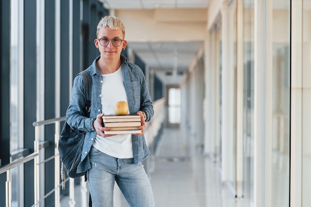 ジーンズの服を着た男性の若い学生は、手にリンゴを持った本を持っている大学の廊下にいます