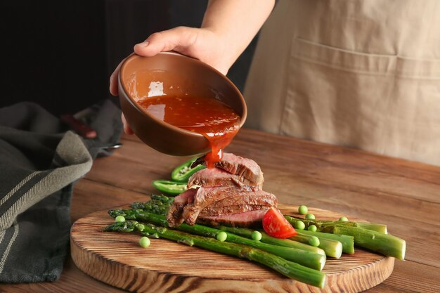 Photo man adding sauce to sliced steak with asparagus on serving board