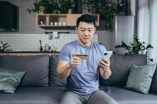 Man alone at home shopping online asian uses phone and shopping app holds credit card sits on sofa at home