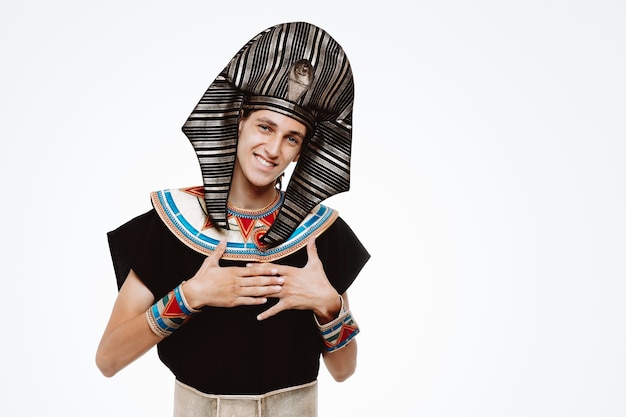 Man in ancient egyptian costume smiling friendly holding hands on his chest feeling thankful on white