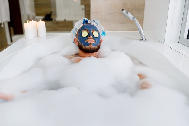 Man applies face mask and relax in bath with foam, morning hygiene. Male person resting in bathroom, skin and body treatments procedures