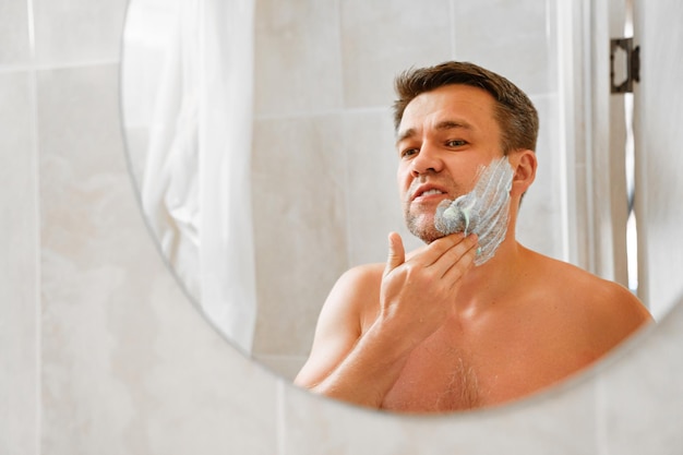 Photo a man applies shaving foam on his face and looks in a round mirror