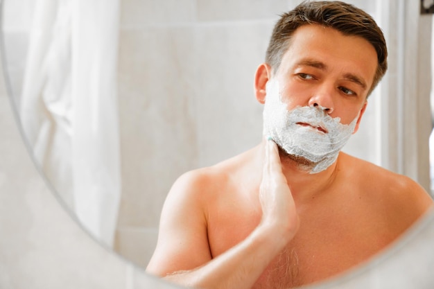 Photo a man applies shaving foam on his face and looks in a round mirror