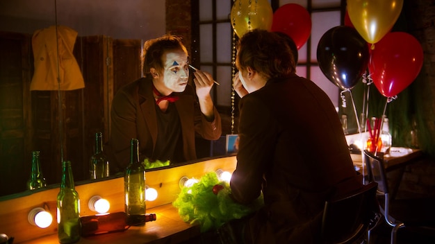 A man applying clown paint on the eyebrows in front of the\
mirror in the dressing room