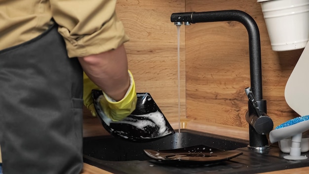 Man in apron washes black plate with sponge and detergent