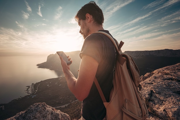 一人旅の山頂でリラックスしたスマートフォンを使用して男のバックパッカー。ライフスタイルアクティブな休暇現代技術ミレニアル世代の概念。素晴らしい夜明け。テキストをモックアップします。