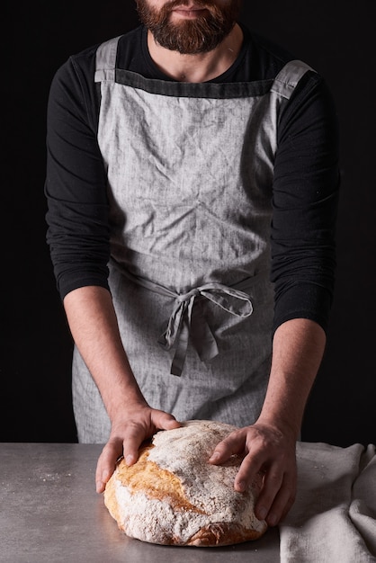Бородатый пекарь в сером фартуке стоит на черном фоне и держит, ломает, отрезает вкусный, хрустящий хлеб, булочки, багет.