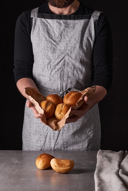 A man baker with a beard in a gray apron stands against a black background and holds, breaks, cuts off delicious, crispy bread, rolls, baguette.