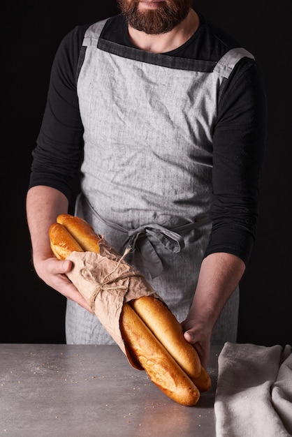 A man baker with a beard in a gray apron stands against a black background and holds, breaks, cuts off delicious, crispy bread, rolls, baguette.