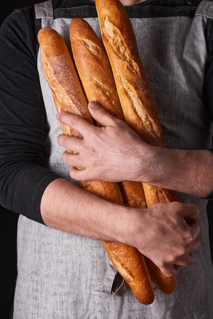 A man baker with a beard in a gray apron stands against a black background and holds, breaks, cuts off delicious, crispy bread, rolls, baguette.