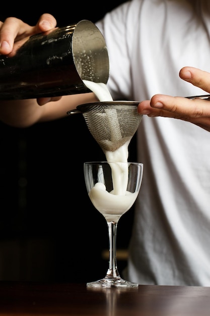 Il barista uomo filtra accuratamente il cocktail pisco sour dalla tazza dello shaker in acciaio nel vetro attraverso il setaccio vista in primo piano