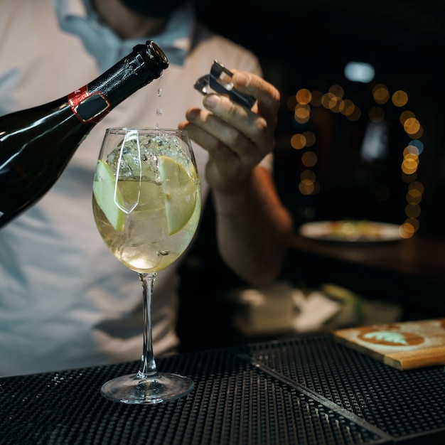 Man bartender pouring sweet champagne into a glass with ice and a slices of apple at a in nightclub party. Lifestyle