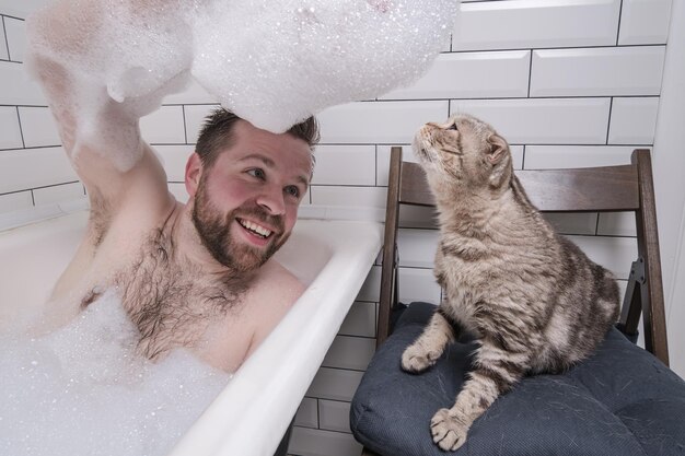 Man bathes in a bathtub he holds on hand a lush soap suds which\
his adored cat is interested in