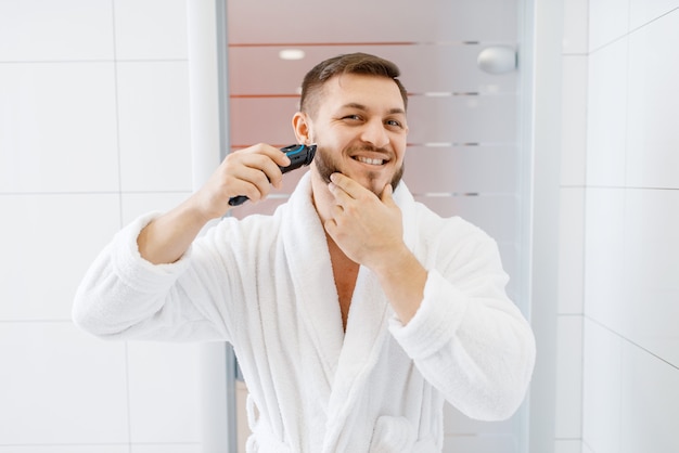 Man in bathrobe shaves his beard with an electric razor in bathroom