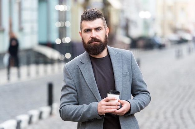 Man bearded hipster drink coffee paper cup. Businessman well groomed enjoy coffee break outdoors urban background. Thoughtful but relaxed. Walk and enjoy fresh hot coffee. Involved in relaxation.