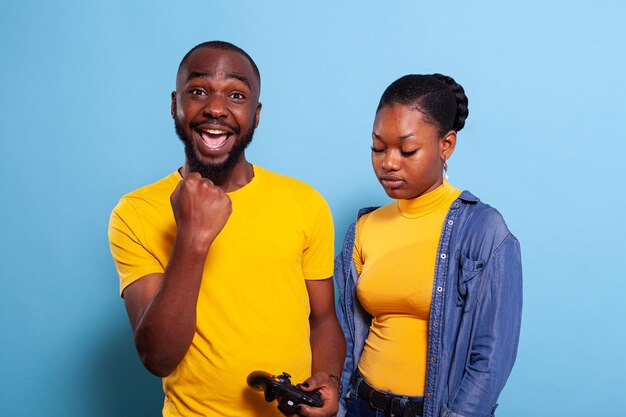 Man beating woman at video games with controller on console over blue background. Boyfriend celebrating win and girlfriend feeling disappointed about losing online gameplay in studio.