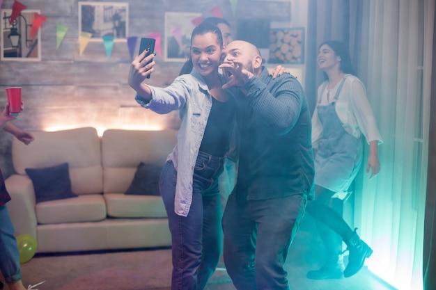 Man and beautiful woman using smartphone taking a selfie while partying with their friends.