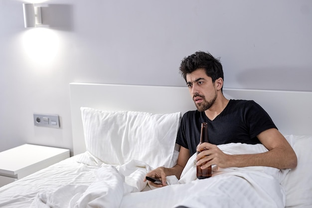 Man in bed with beer watching tv. alcoholism from boredom\
during covid-19 coronavirus quarantine epidemic. stay at home\
concept. alcoholic male alone at home, in bright bedroom